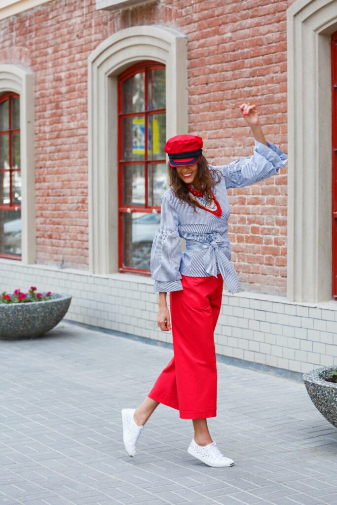 Stylish brunette girl dressed in a striped blouse, red wide pants and a red cap poses in the city